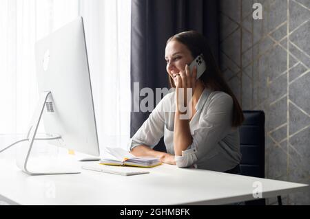 Charming young woman has phone conversation, a business lady relaxed sits at the desk in front of trendy computer, laugh and chatting on the smartphone while take a break from remote working Stock Photo