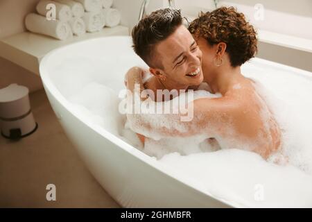 Smiling young queer couple enjoying a bubble bath together. Romantic young couple having fun together in the bathtub at home. Young LGBTQ+ couple spen