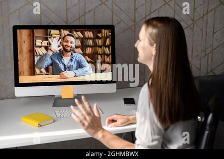 A friendly female office worker in casual wear, sitting at the desk in the home office, waving at the laptop, greeting participants of online conference, saying hello to Indian male coworker Stock Photo