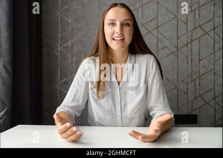 Smiling brunette young woman holding webcam digital conversation, looks at the camera and talking with gestures. Female chatting on the distance. Virtual event concept. Stock Photo