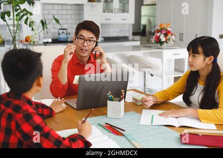 Asian father working on laptop at table with son and daughter doing school work Stock Photo