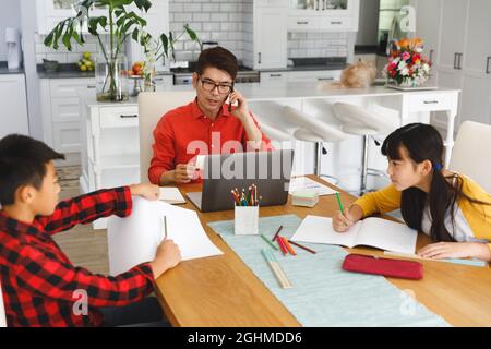 Asian father working on laptop at table with son and daughter doing school work Stock Photo