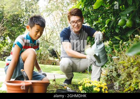 Happy asian father and son smiling, wearing gloves and watering plants together in garden Stock Photo