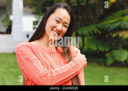 Portrait of happy asian woman smiling in garden outside family home wearing pink sweater Stock Photo