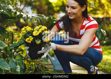 Smiling caucasian woman working in garden, holding flowers and wearing gloves Stock Photo