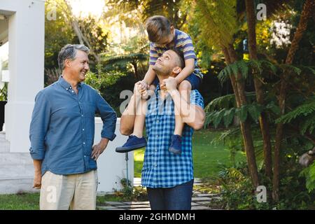 Portrait of smiling caucasian grandfather and adult son with grandson on his shoulders in garden Stock Photo