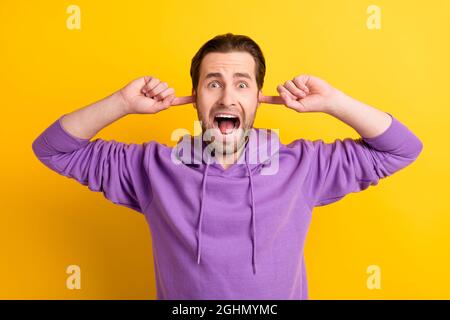 Photo of crazy mad tired annoyed guy finger close ears scream wear violet hoodie isolated yellow color background Stock Photo