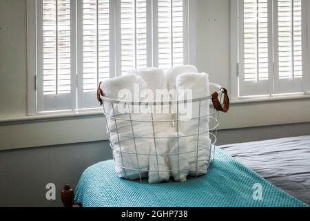 A basket of organized clean rolled white towels for guests sitting on a bed in a guest bedroom Stock Photo