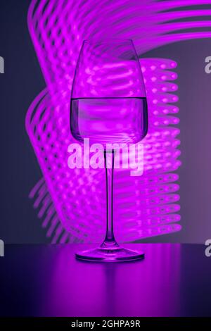 Elegant crystal glass filled with pure water on a purple background painted with colorful light. Light reflections and refractions, light streaks, par Stock Photo