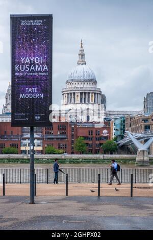 London, United Kingdom - July 30, 2021: Two people walk along the bank of river Thames outside Tate Modern gallery with St Paul's Cathedral and the Mi Stock Photo