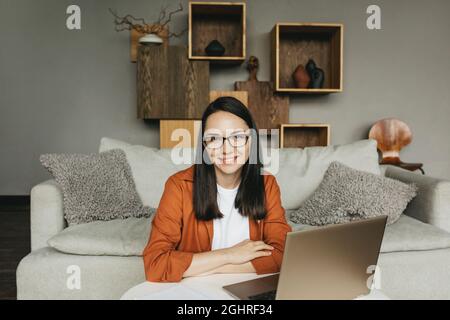 A beautiful European young woman in casual clothes, using a laptop from home. Conducts a video call, communicates online with colleagues, sends and responds to emails, develops software while sitting at home on the couch. A smart woman works remotely Stock Photo