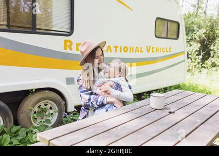 Beautiful millennial caucasian mother hugging daughter near camper van in forest. Young stylish woman in shirt and cowboy hat enjoying life and having Stock Photo