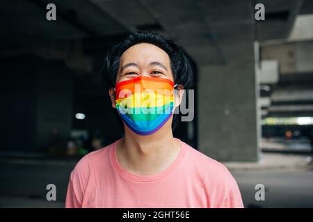 Young happy Asian man wearing a rainbow patterned mask representing LGBT support and pride during Covid-19 or Coronavirus Stock Photo