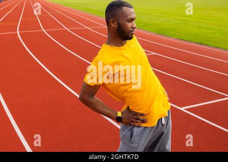 Sportive young adult man standing at stadium track, warming up before running. African american male is working out, stretching back, training
