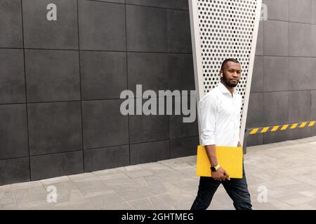 Young adult male student or freelancer in stylish clothes standing with yellow laptop outdoors, walking to the office for business meeting. Serious Stock Photo