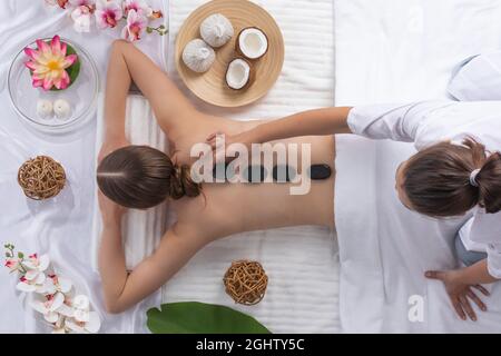 Spa stone massage, beautiful woman getting hot stones massage, beauty treatments concept. Top view. Orchid and lotus flowers coconut and herb pouches Stock Photo