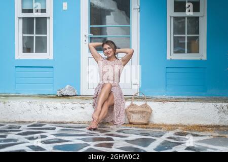 Pretty young caucasian woman with blond hair sitting near cozy coffee shop with blue walls and smiling. Holding her hair. Woven summer bag Stock Photo