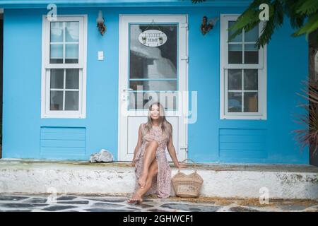 Pretty young caucasian woman with blond hair sitting near cozy coffee shop with blue walls and smiling with the teeth. Laughing girl. Woven straw bag Stock Photo