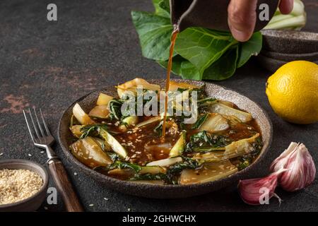 Man hand pouring soy sauce in bok choy or chinese cabbage with sesame seeds on dark background. Asian chinese food. Healthy vegan or vegetarian food Stock Photo