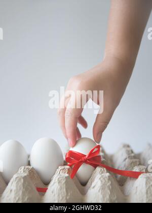 Woman's hand takes white egg with red gift ribbon in an egg carton on a white background. Copy space. Place for text. Vertical. Stock Photo