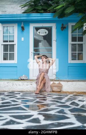 Pretty young caucasian woman with blond hair sitting near cozy coffee shop with blue walls and smiling with the teeth. Holding her hair. Woven bag Stock Photo