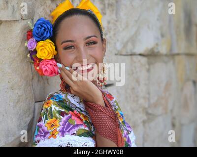 Young beautiful Mexican woman with makeup wears traditional Mayan Yucatecan folkloric dress with flowers in her hair and smiles for the camera. Stock Photo