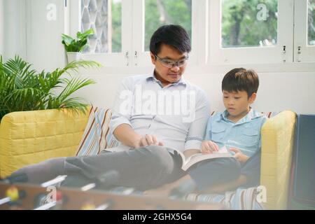 Father and son sitting on a sofa reading a book together Stock Photo