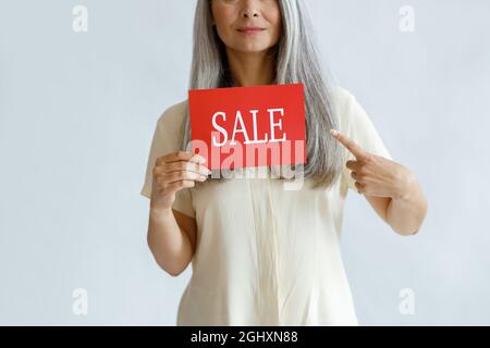 Lady with long grey hair holds red Sale sign pointing by finger on light background Stock Photo