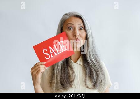 Funny middle aged Asian woman shows red Sale sign grimacing on light background Stock Photo