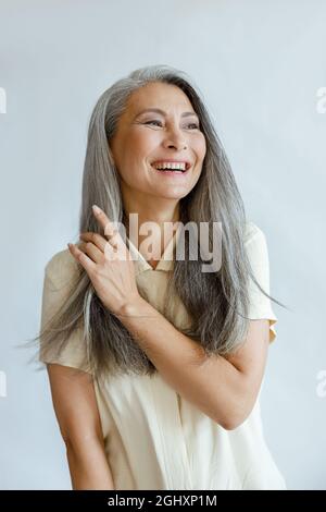 Middle aged Asian woman touches natural hoary hair on light grey background Stock Photo