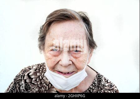Close-up of Woman over 80 years of age smiling with protective mask down. Face with age wrinkles and blue-gray eyes. Wearing a printed shirt. White ba Stock Photo