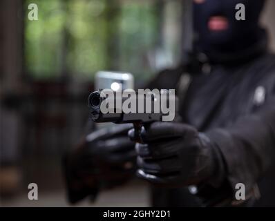 Videotaping your death concept. Blur terrorist in black balaclava holding pistol in leather gloved one hand and mobile in the other. Armed man killer Stock Photo