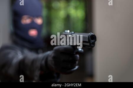Terrorist in black balaclava, holding a gun in gloved hand. Blur man aiming with pistol background. Hooded killer armed murderer robbery. Death is nea Stock Photo