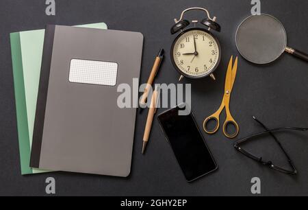 School teacher or student office desk flat lay, workspace. Notebooks, mobile phone stationery and alarm clock on black color background, top view. Stock Photo
