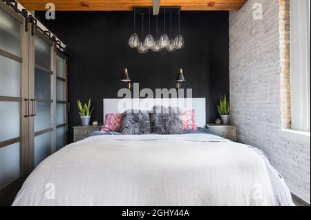 A bedroom in a loft condo with an exposed wood ceiling, glass sliding doors to a closet, black accent wall, and a fancy glass light hanging above. Stock Photo