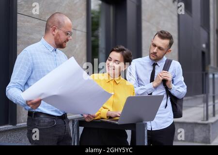 Business people, designers or architecture managers in casual clothes discussing project or presentation outdoors near modern building using laptop. Discussing project concept. High quality image Stock Photo