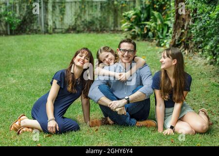 A family of a mother and father and two daughters sitting outside in the grass of their backyard witht the youngest daughter hugging dad from behind Stock Photo