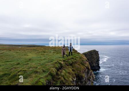 Young man and woman wearing jacket standing at the edge of Moher cliff in Ireland while travelling and enjoying the nature during day under cloudy sky Stock Photo