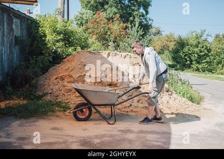 A 30-year-old caucasian man pours sand from a large pile of sand onto a garden wheelbarrow Stock Photo