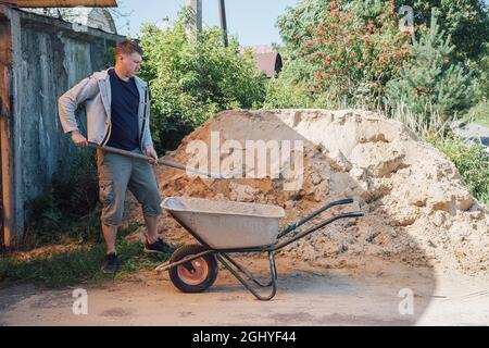 A 30-year-old caucasian man pours sand from a large pile of sand onto a garden wheelbarrow Stock Photo