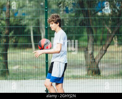 Cute teenager plays basketball at city playground. A boy holds basketball ball in his hands outside. Active life, hobby, sports for children Stock Photo