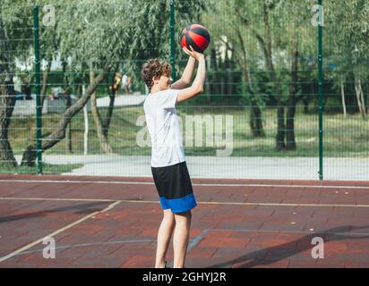 Cute teenager plays basketball at city playground. A boy holds basketball ball in his hands outside. Active life, hobby, sports for children Stock Photo