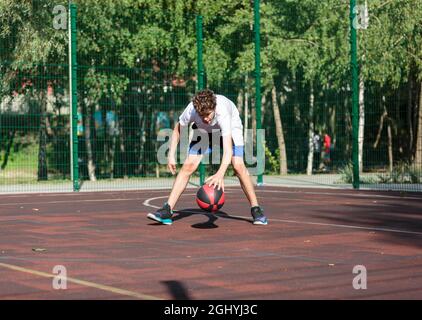 Cute teenager plays basketball at city playground. A boy holds basketball ball in his hands outside. Active life, hobby, sports for children Stock Photo