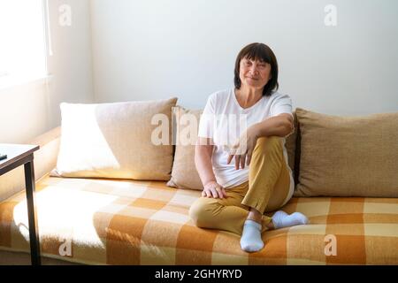 Portrait shot in head of peaceful calm middle aged woman relaxing on cozy sofa alone at home. Nice happy 60s old mature grandmother relaxing on living room couch posing for photo. Stock Photo