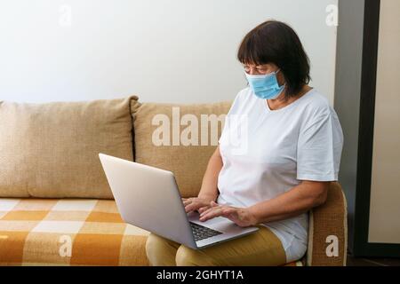 Serious woman in protective mask uses laptop, checks email on internet while sitting on couch, looking for friends on social networks or working on the computer, blogging at home Stock Photo