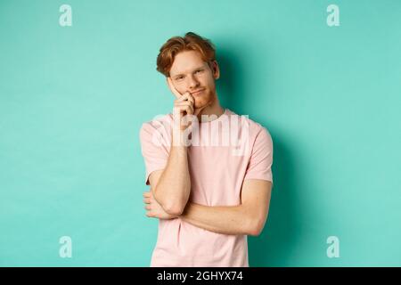 Bored handsome guy with red hair leaning on hand, looking tired and unamused at camera, standing in t-shirt against turquoise background Stock Photo