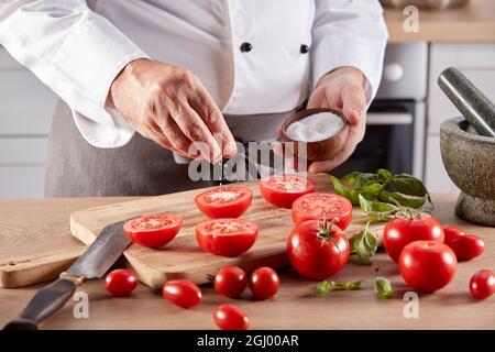 Crop faceless male chef sprinkling salt on halves of ripe fresh tomatoes while preparing food at wooden table in kitchen Stock Photo