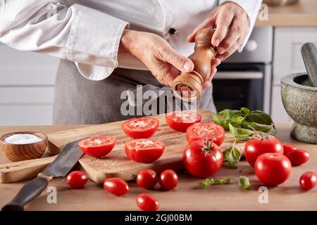 Unrecognizable crop male chef with pepper mill sprinkling spices on fresh tomatoes halves while preparing food at table in kitchen Stock Photo