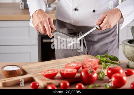 Anonymous crop male chef sharpening blade of knife while standing at wooden table with tomatoes before cooking in kitchen Stock Photo