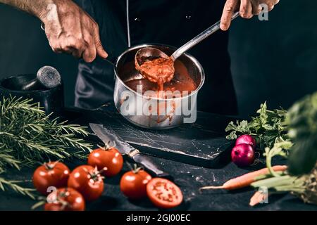 Crop unrecognizable male cook stirring tasty tomato soup with ladle while preparing healthy lunch at table with assorted ingredients in kitchen Stock Photo